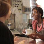 Eco-Friendly Shopping - Smiling African American waitress wearing casual t shirt and apron standing at counter in cozy coffee shop with female customer while serving hot drink and looking at each other